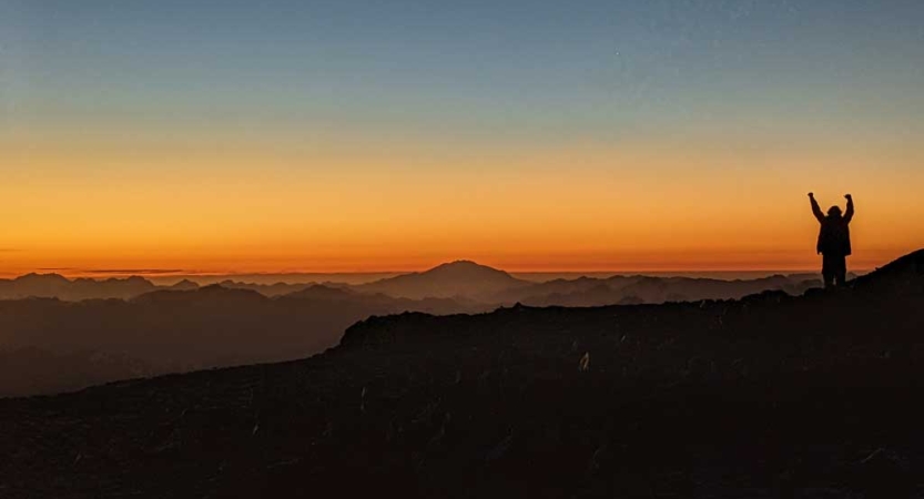 A person stands on a mountainous overlook and raises their hands into the air. The sky appears in shades or orange, yellow and blue, as the sun is either rising or setting. 